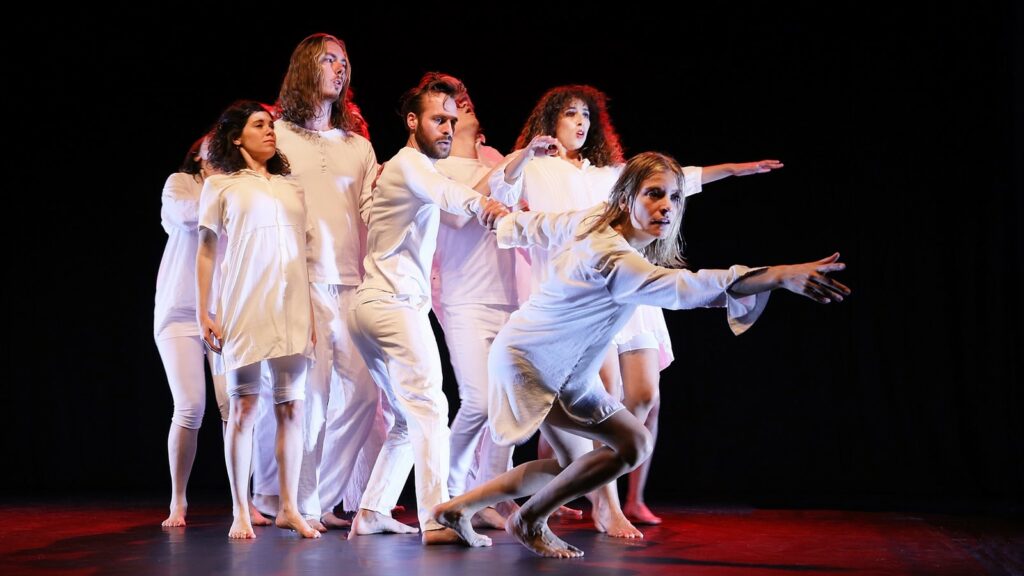 Sieben tanzende Sängerinnen und Sänger in weißer Kleidung bilden eine Gruppe, aus der eine Persone nach vorne strebt