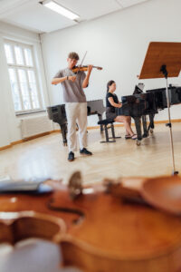 Violine liegt im Vordergrund; hinten ein junger Mann, der Geige spielt und von einer Pianistin in dunkler Kleidung begleitet wird.